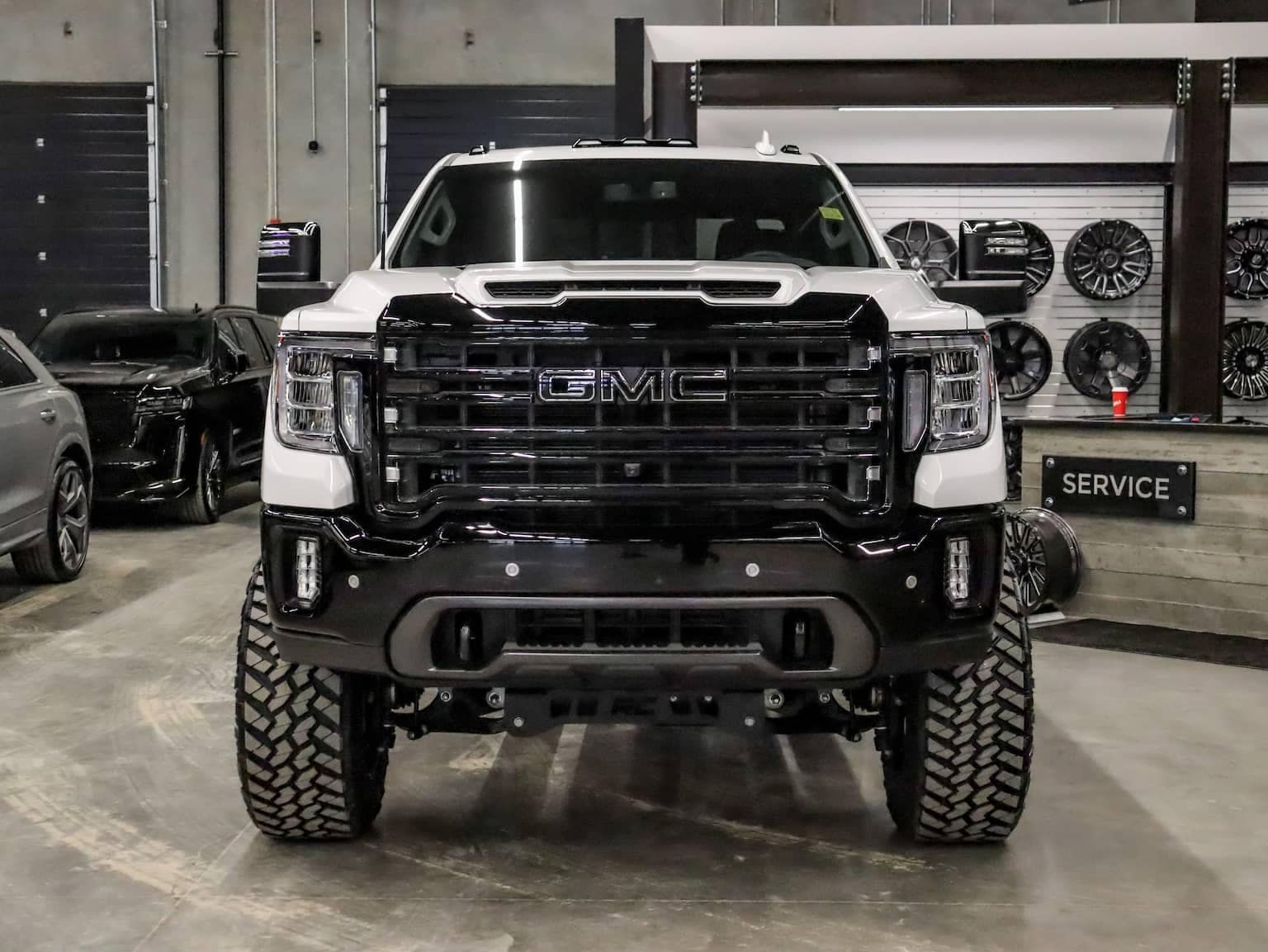 GMC truck front view showcasing a robust design with bold black grille and oversized tires, set in a vehicle service center environment with various car models in the background.