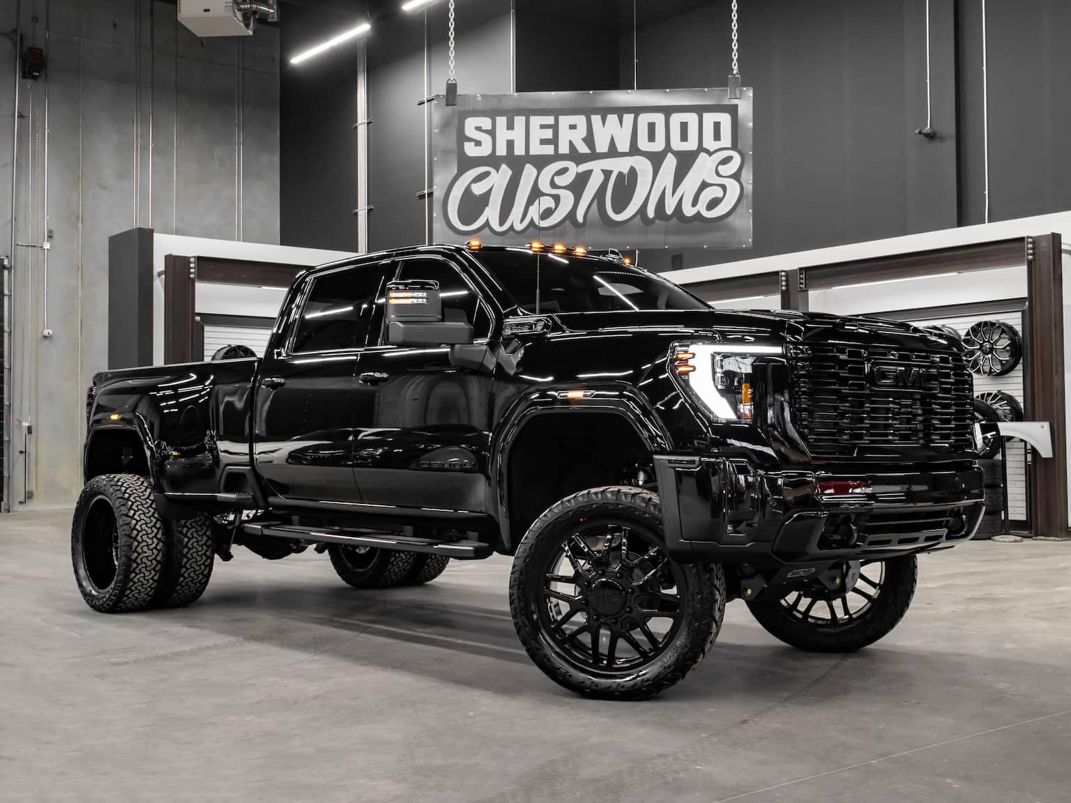 Custom black GMC pickup truck showcased in a contemporary showroom, featuring large wheels and a bold design, highlighting Sherwood Customs branding in the background. Ideal for automotive enthusiasts and truck customization inspiration.