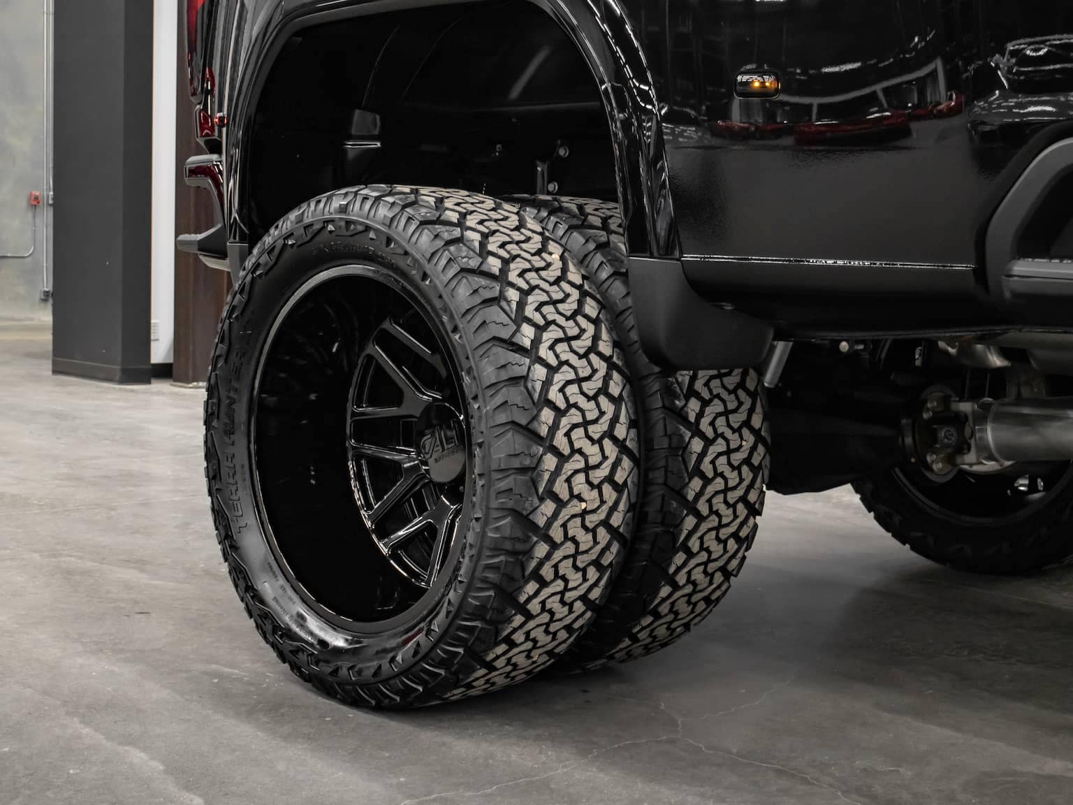 Close-up view of an off-road truck displaying oversized, all-terrain tires mounted on stylish black alloy wheels. The image showcases the rugged tread design, emphasizing durability and performance in various terrains, set against an industrial backdrop.