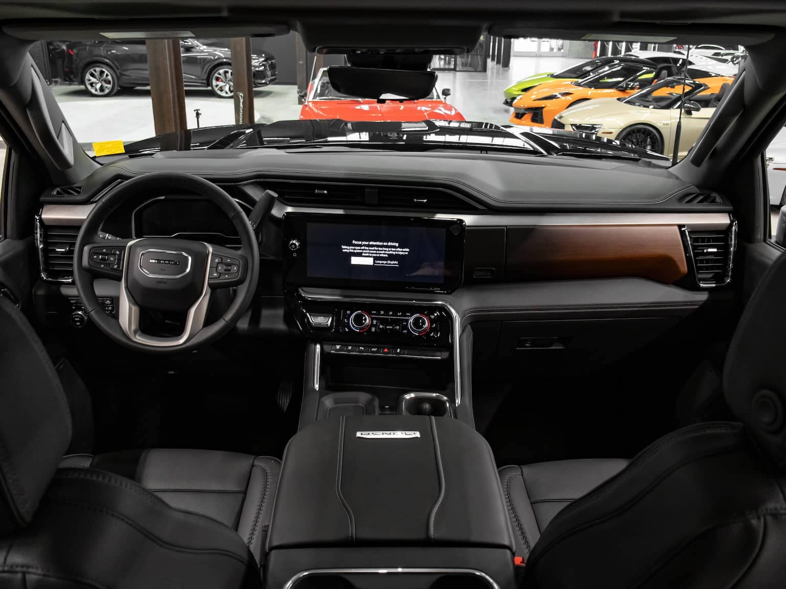 Interior view of a luxury SUV showcasing a modern dashboard with a touchscreen display, leather seats, and a sleek design. Multiple sports cars are visible in the background, highlighting the upscale setting.