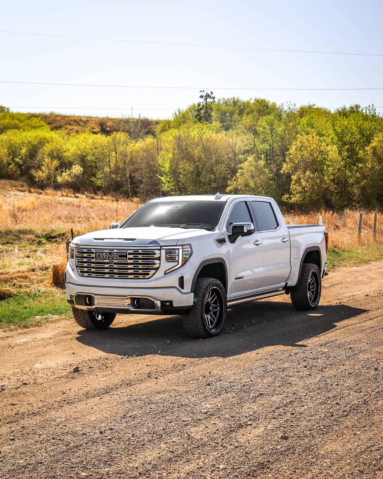 White GMC truck parked on a dirt road surrounded by greenery, showcasing its sleek design and rugged tires, ideal for off-road driving.