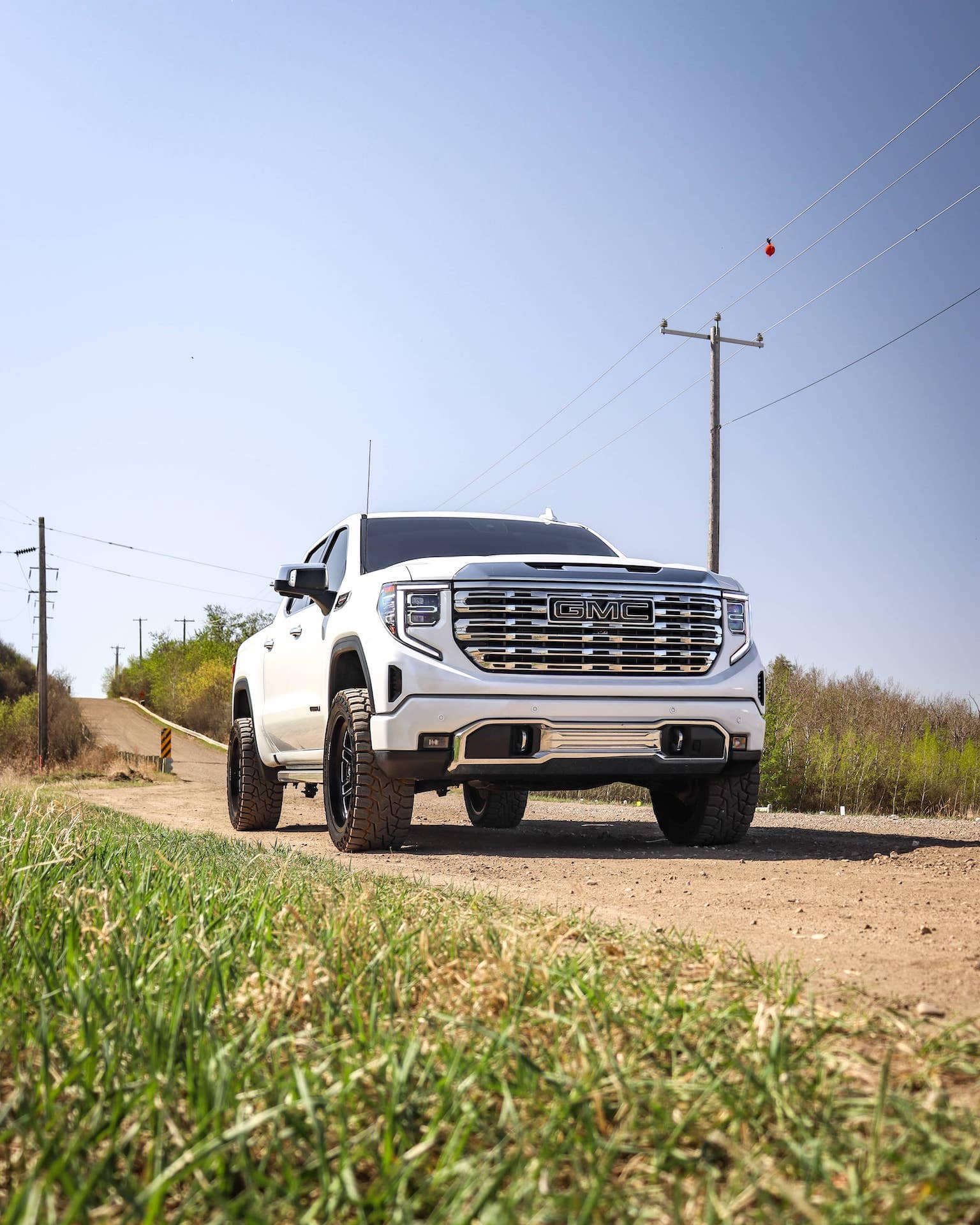 White GMC truck parked on a dirt road surrounded by greenery and power lines under a clear blue sky. The vehicle showcases a modern design with prominent front grille and off-road tires, highlighting its rugged appeal.