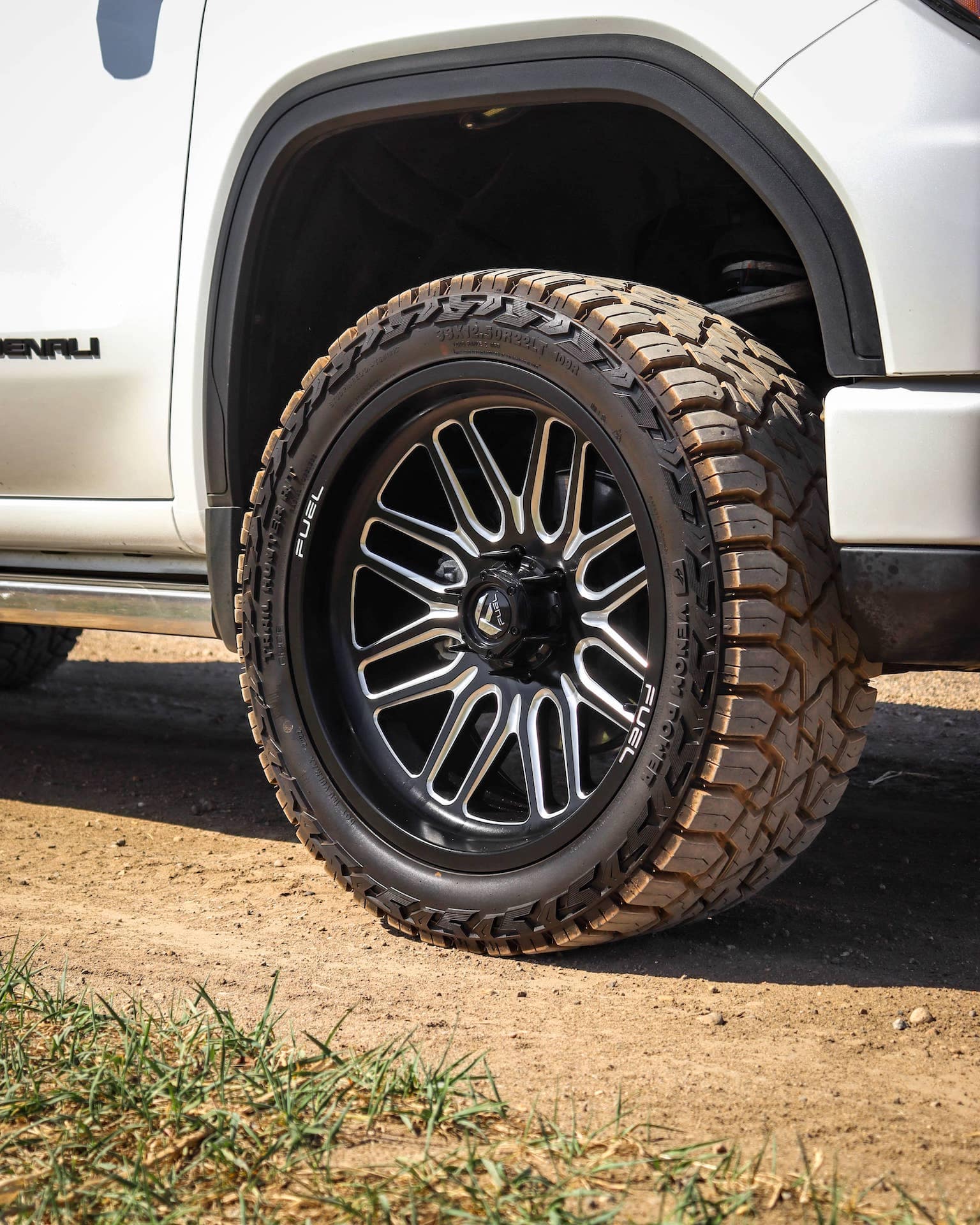 Close-up of a black and chrome alloy wheel with a rugged all-terrain tire mounted on a white SUV, showcasing the vehicle's stance against a dirt background. The image highlights the wheel design and tire tread pattern, emphasizing durability and style for off-road driving.
