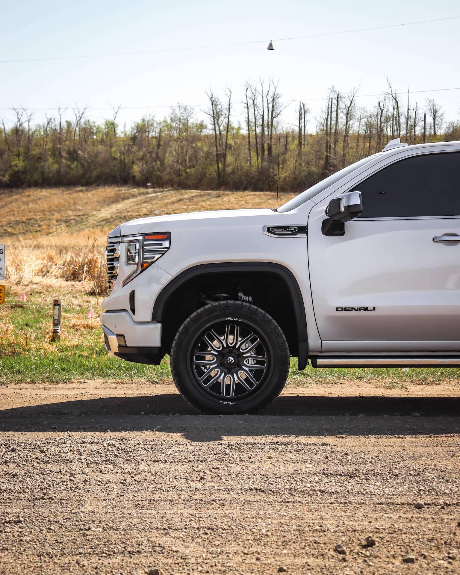 Side view of a white GMC Denali truck parked on a gravel surface, showcasing its sleek design, custom wheels, and off-road tires, with a natural background of sparse trees and open field.