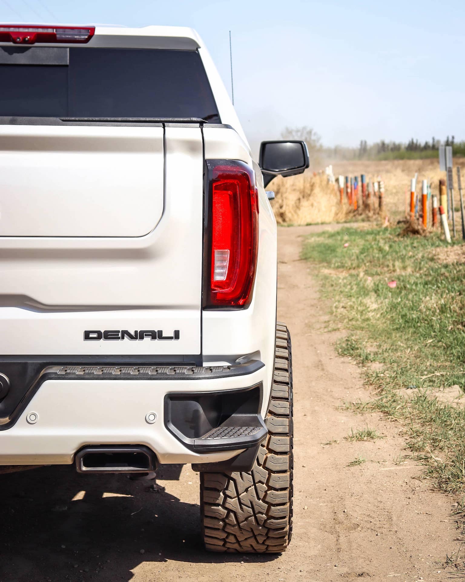 Close-up of the rear of a white GMC Denali pickup truck parked on a dirt road, showcasing its rugged tires and modern tailgate design against a backdrop of open fields.