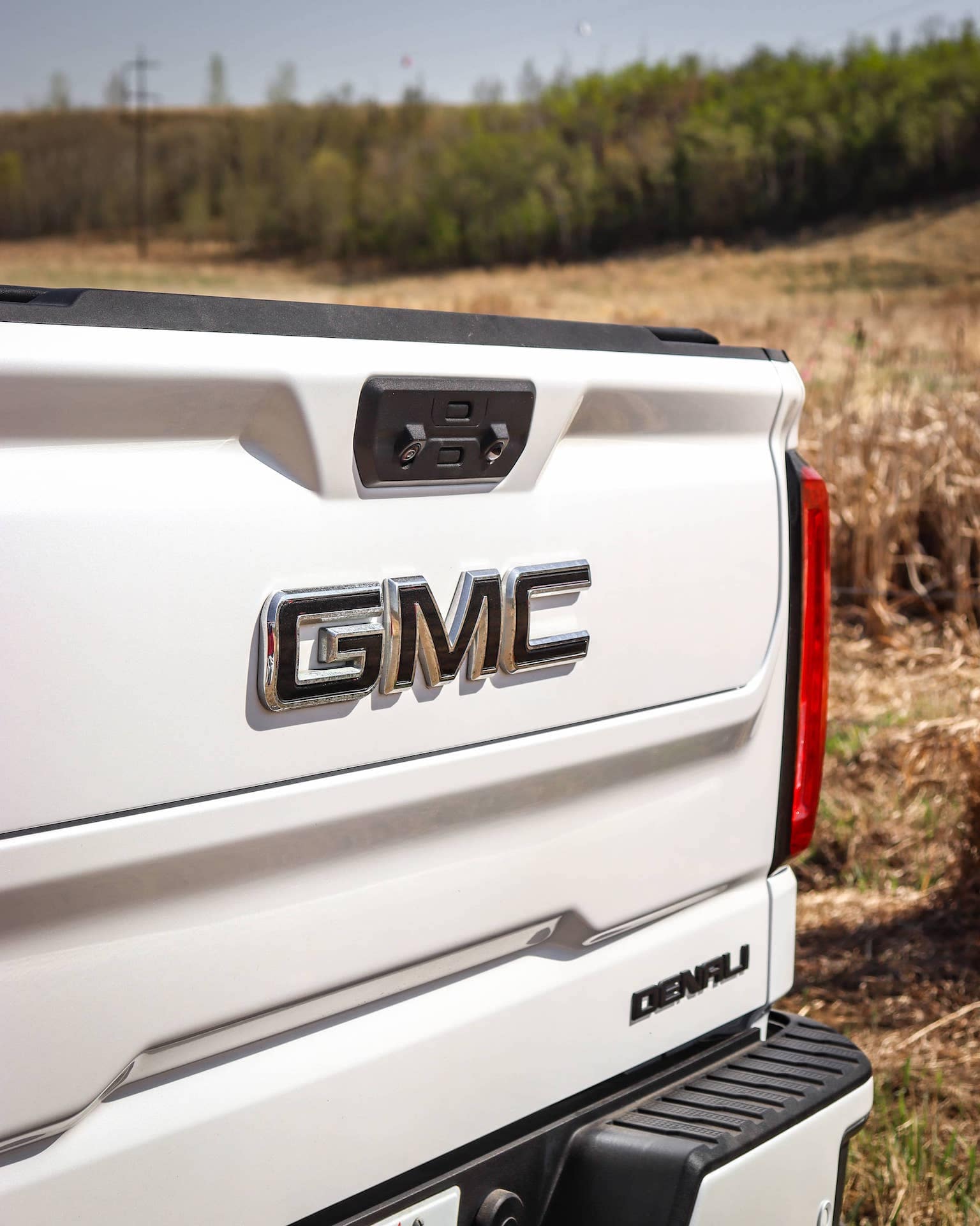 Close-up of the rear of a white GMC Denali truck, featuring the prominent GMC logo, tailgate handle, and a scenic background of green trees and open fields. Perfect for showcasing GMC's rugged design and premium features.