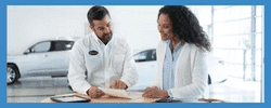 Car dealership sales representative discussing vehicle options with a female customer at a table. The background features cars in a well-lit showroom, highlighting a professional and welcoming atmosphere.