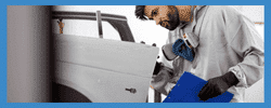 Auto body technician inspecting vehicle door during repair process, wearing safety gloves and a mask, with a clipboard in hand.
