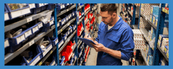 Warehouse employee checking inventory on a clipboard while standing among organized shelving units filled with various tools and parts.