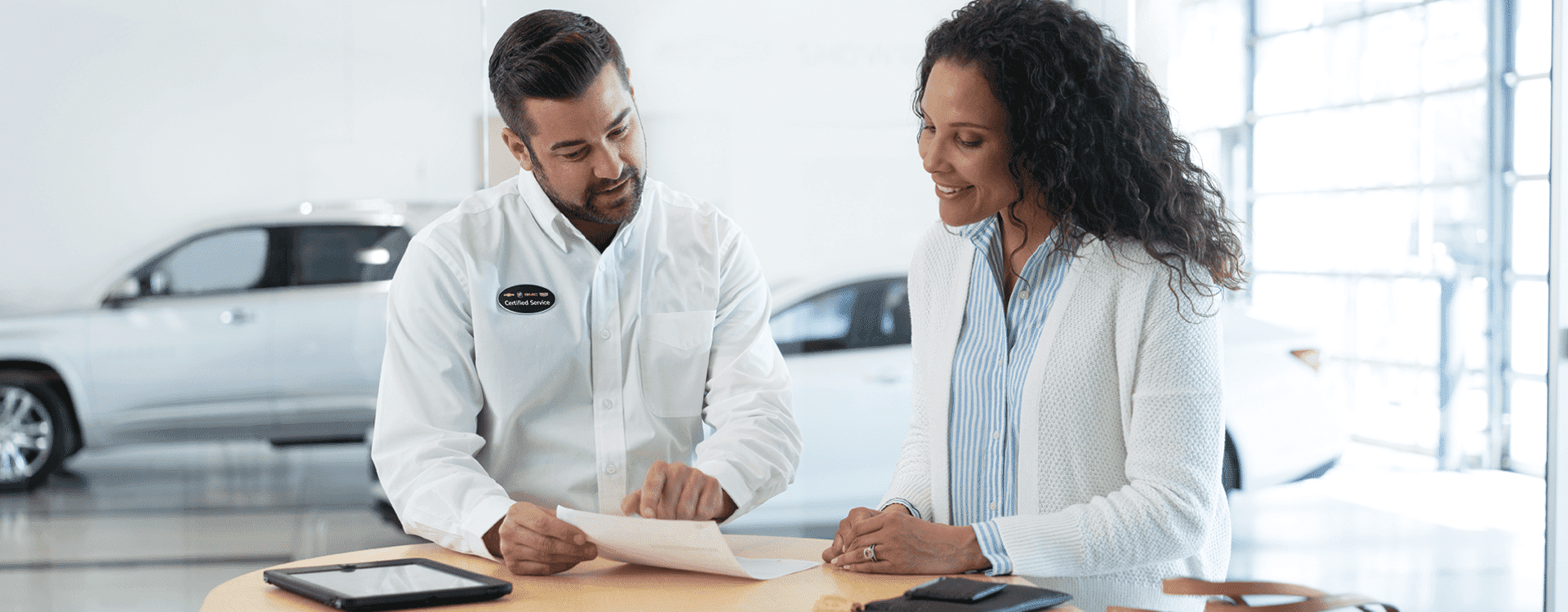 A car dealership employee discusses service options with a smiling customer at a reception desk, surrounded by a bright showroom with vehicle displays in the background.