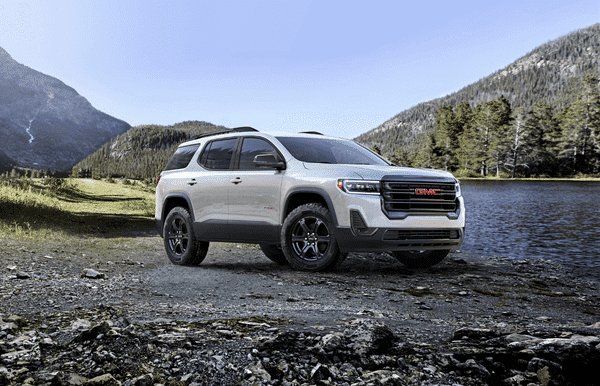 GMC Acadia SUV parked by a serene lake surrounded by mountains and trees, showcasing its sleek design and off-road capability against a natural landscape.