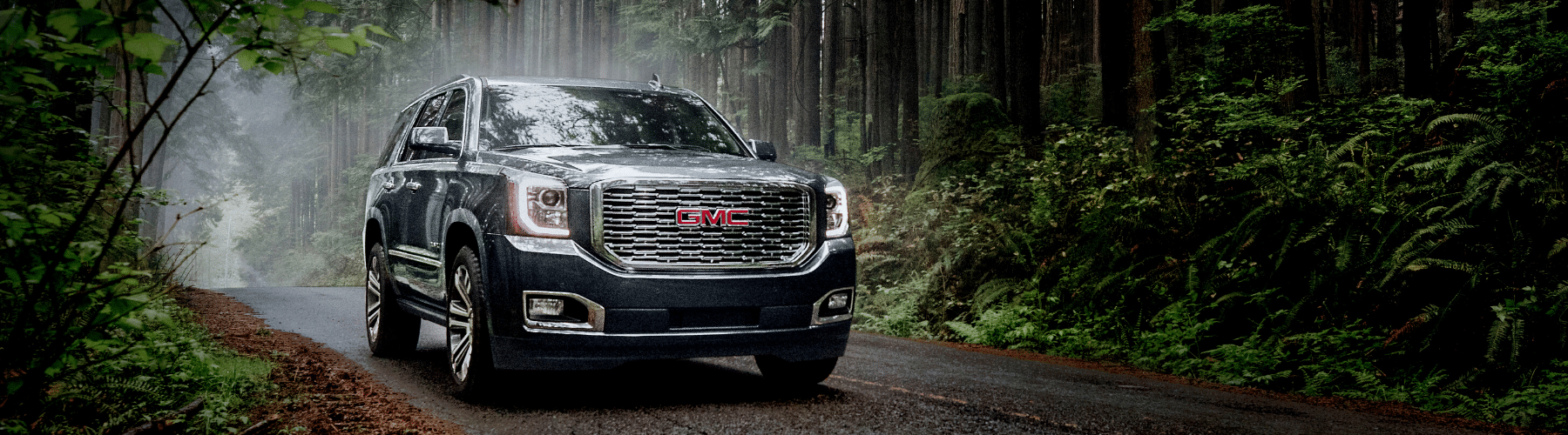 GMC SUV driving through a misty forested road, showcasing its rugged design and off-road capability amidst lush greenery.