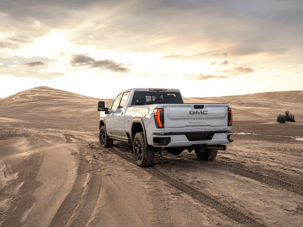 GMC truck parked on sandy terrain with tire tracks, showcasing off-road capabilities against a scenic sunset backdrop.