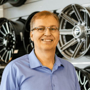 Smiling man in a blue shirt standing in front of a display of car wheels, showcasing expertise in automobile accessories.