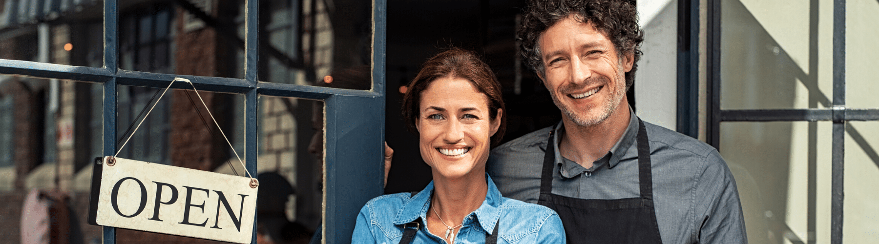 Smiling business owners standing in front of their shop, displaying an "Open" sign. The image captures a welcoming atmosphere, highlighting the couple's friendly demeanor and the inviting entrance of their retail location.