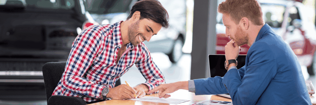 Two men engaged in a business meeting at a car dealership, discussing documents and finalizing a car purchase. One man is signing a document while the other points to details on a brochure, with cars visible in the background.