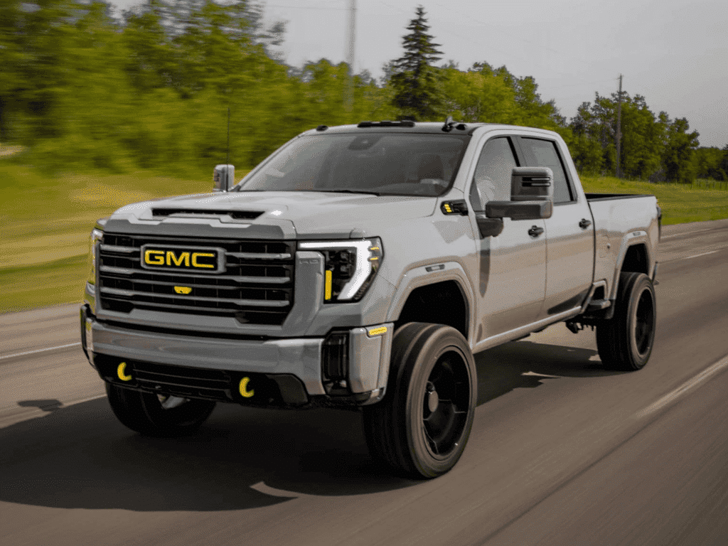 GMC pickup truck driving on a rural road, showcasing a sleek silver exterior, bold front grille with GMC branding, and distinctive black wheels, emphasizing its powerful and rugged design.