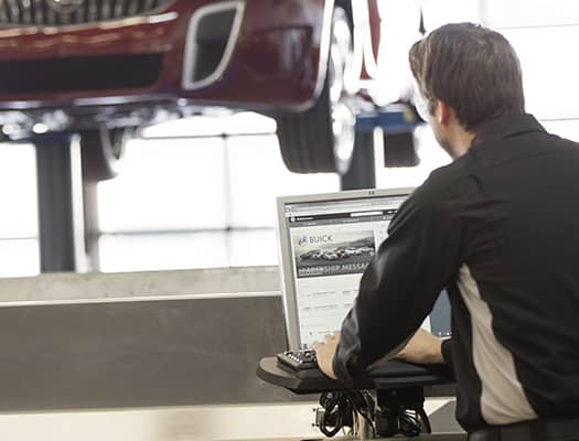 Automotive technician working on a computer in a service center with a Buick vehicle elevated in the background, illustrating vehicle maintenance and diagnostics.