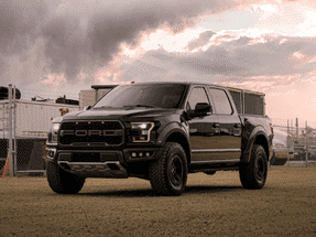 Black Ford F-150 Raptor truck parked on a gravel lot with industrial background under a dramatic sky.