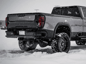 Gray pickup truck with large tires parked in snowy environment, showcasing its rugged design and off-road capabilities. Snow is falling, highlighting winter conditions.