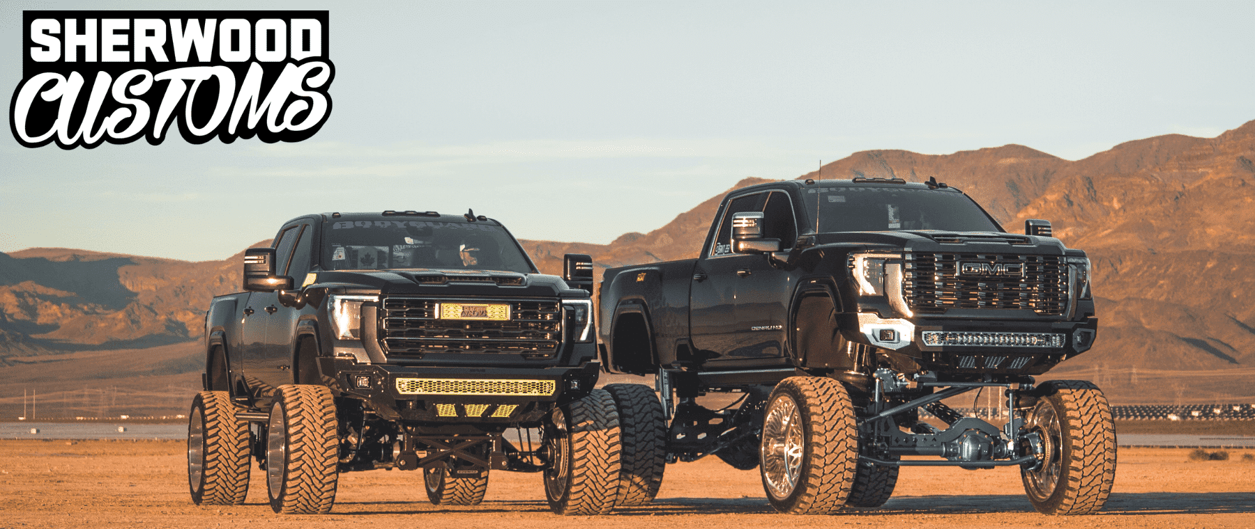 Two customized lifted trucks from Sherwood Customs are showcased in a desert landscape, highlighting their impressive height and rugged design. The trucks feature oversized tires, aggressive styling, and unique details, set against a backdrop of mountains and clear skies, illustrating the brand's commitment to high-performance vehicle enhancements.