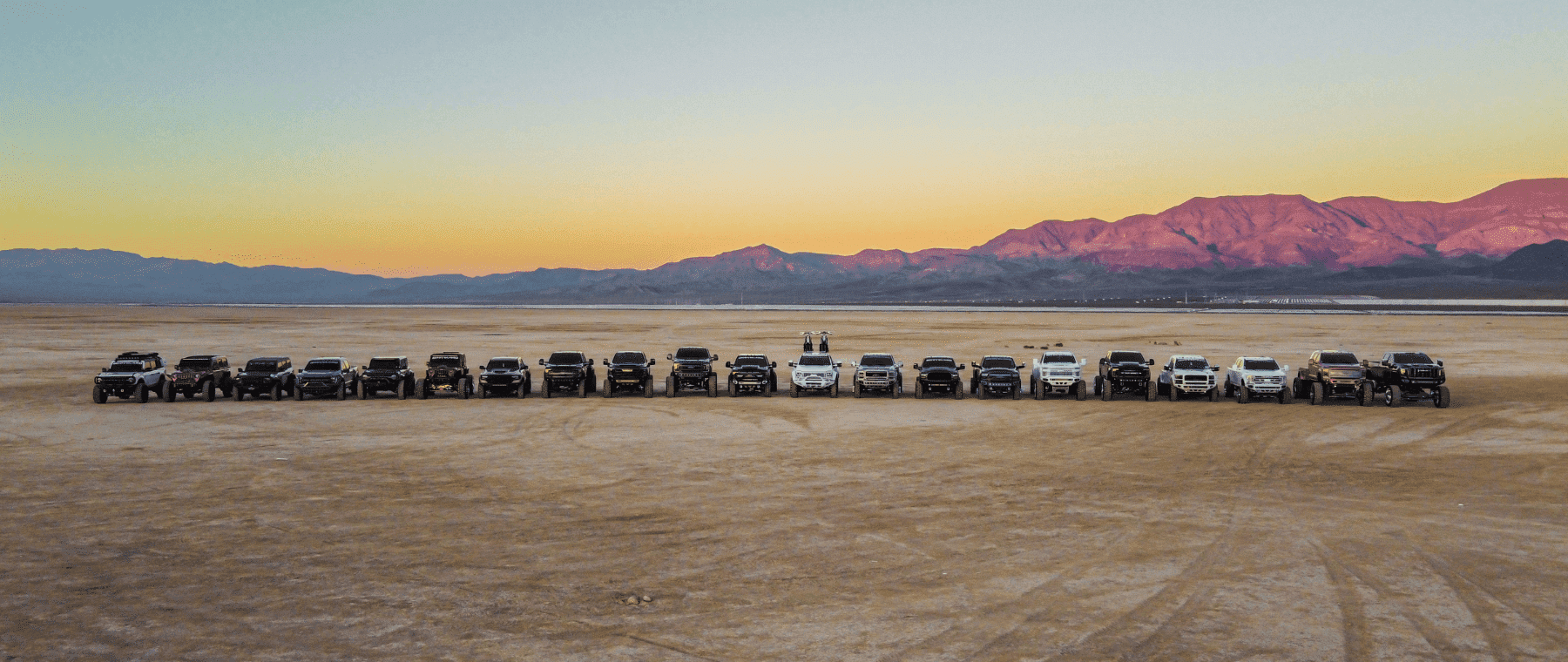 Aerial view of a lineup of off-road trucks arranged on a vast desert landscape during sunset, with mountains in the background. The diverse vehicles, including various makes and models, showcase their rugged design against a colorful sky, emphasizing adventure and exploration.