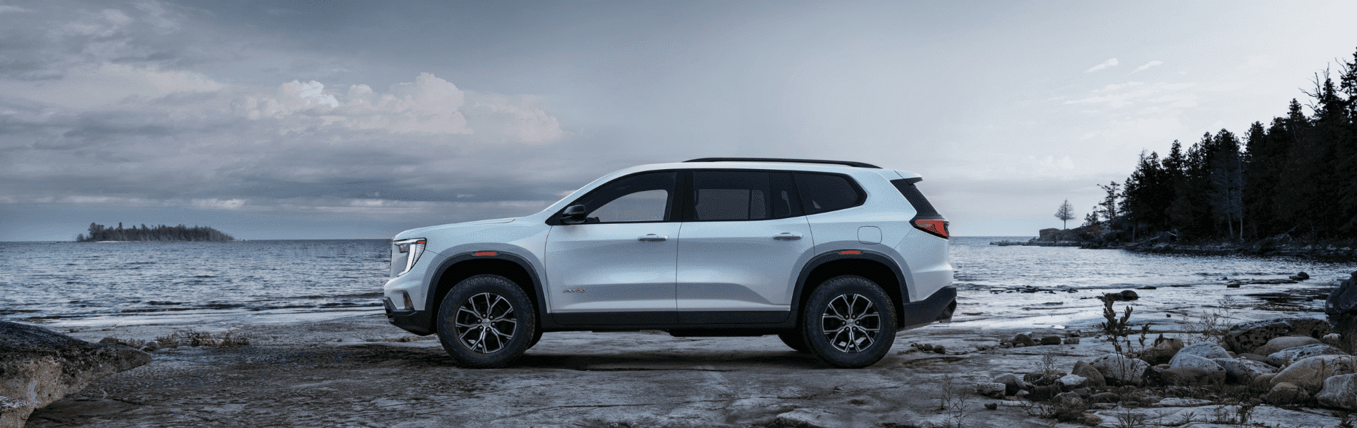 Side view of a white SUV parked on rocky shore near a lake, surrounded by trees and a cloudy sky, showcasing off-road capabilities and an adventurous lifestyle.