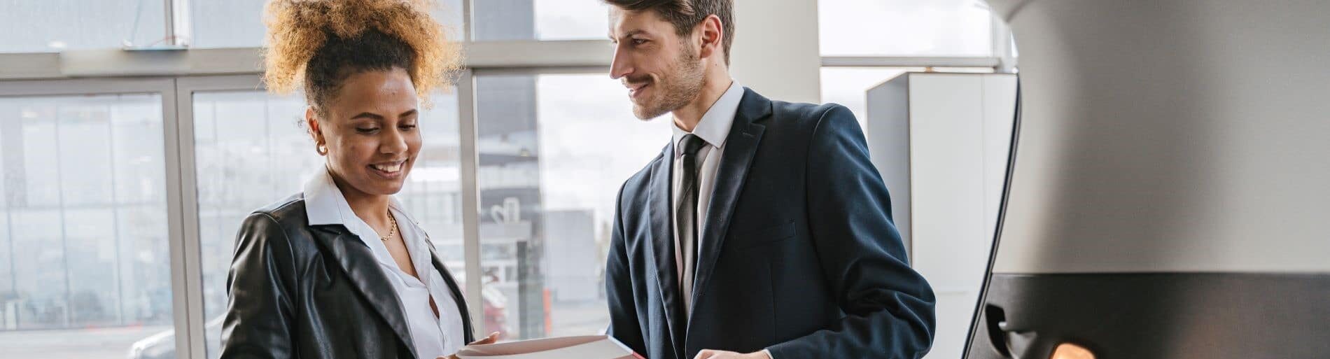 Two professionals, a man in a suit and a woman in a leather jacket, engage in a discussion while reviewing documents in a modern office setting, emphasizing teamwork and collaboration in the workplace.