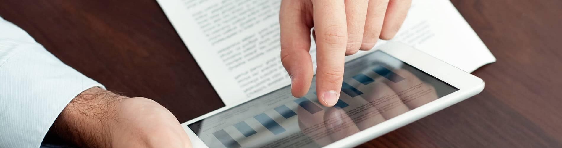 Close-up of a hand interacting with a tablet screen displaying data, with a document partially visible in the background on a wooden desk.