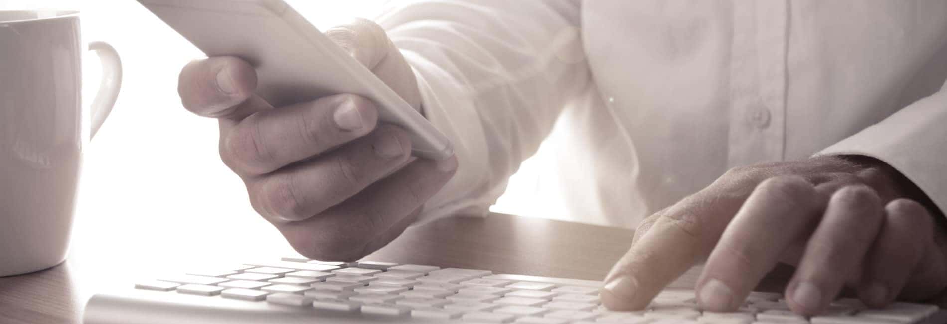 Person using a smartphone while typing on a keyboard, with a coffee cup nearby, illustrating multitasking in a modern workspace.