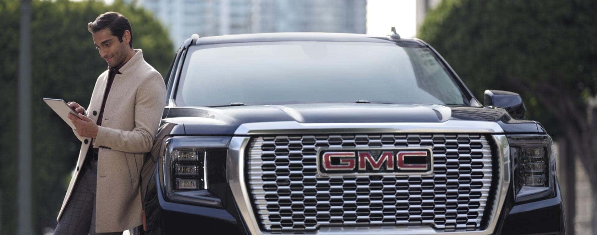 Businessman using a tablet while leaning against a GMC truck, showcasing modern lifestyle and automotive style in an urban setting.