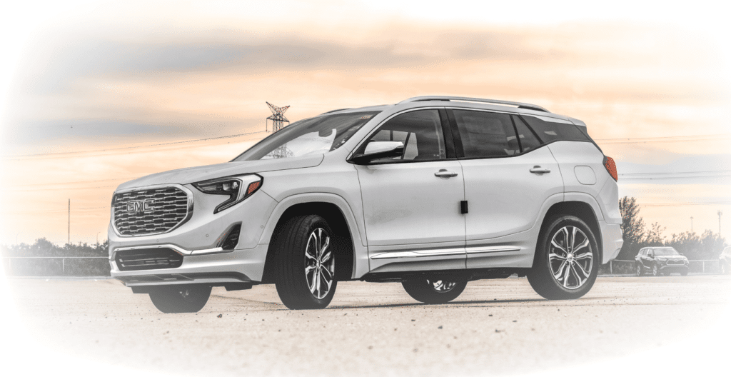 White GMC Terrain SUV parked outdoors with a scenic sunset background, showcasing its sleek design and alloy wheels. Ideal for families and outdoor adventures, blending style with functionality.