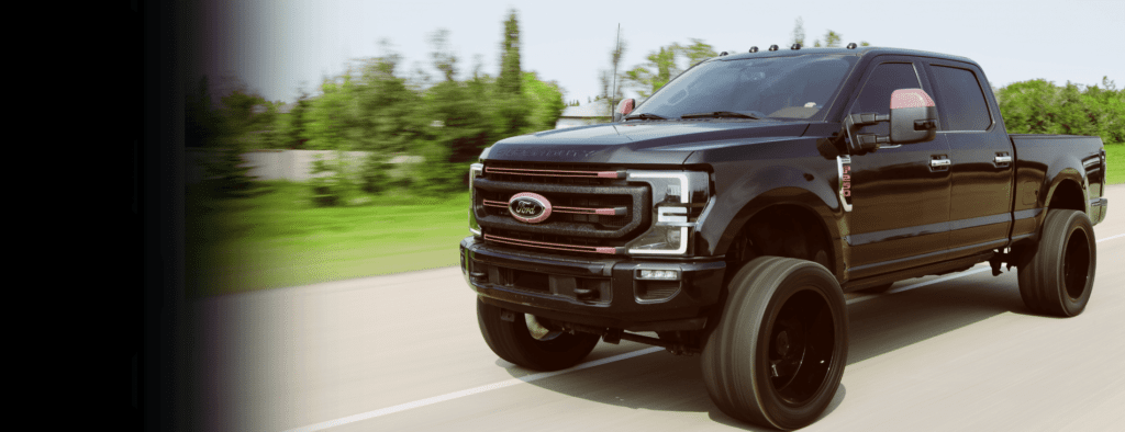 Black Ford Super Duty pickup truck driving on a rural road, showcasing a sleek design with large custom wheels and a rugged appearance, surrounded by greenery.